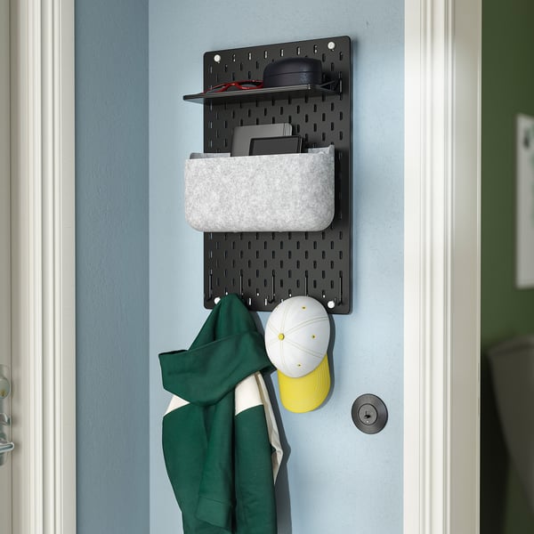 Wall-mounted SKÅDIS organiser with hooks and pockets holding hats and a jacket, offering tidy storage.