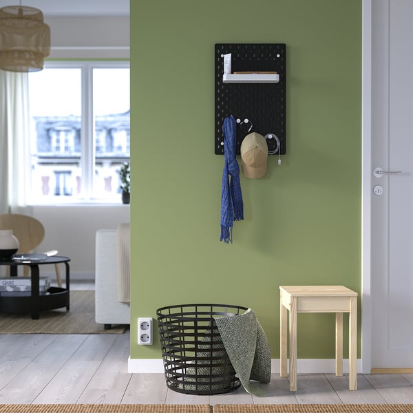 Black SKÅDIS pegboard with accessories on green wall, next to white door and wooden stool, displaying a hat, scarf, and basket.