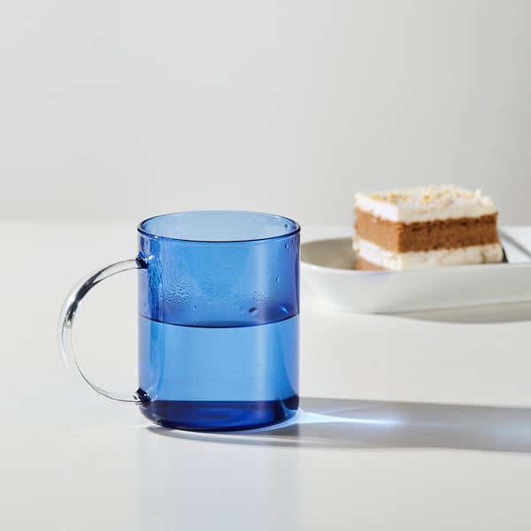 Blue glass mug with handle, half-filled with water, sits near a slice of cake on a white tray.