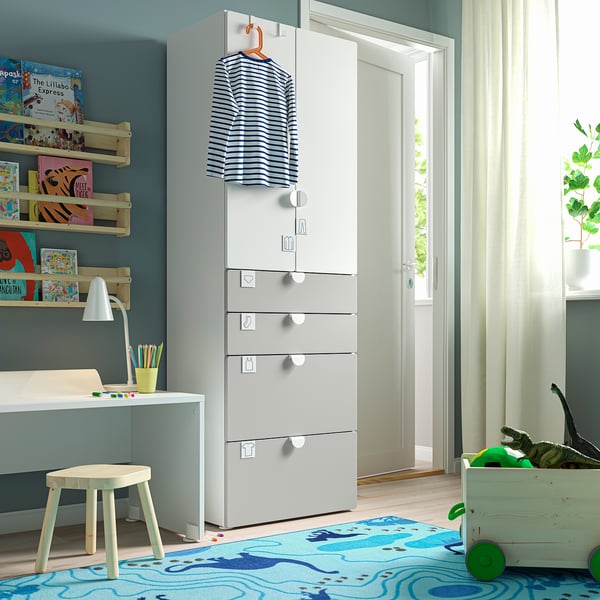 Small childrens room with blue walls, white furniture. Wardrobe with blue shirt, shelves with books, small desk, and toys.