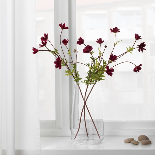 A clear vase holds dark red artificial flowers with green stems, displayed on a white ledge against a window.