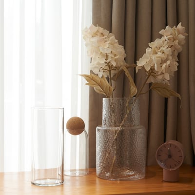 Clear vases and artificial flowers with beige petals and brown stems on a wooden table next to a window with curtains.