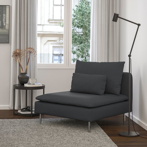 SÖDERHAMN chair in dark grey, sleek & low, placed near a large window with curtains, beside a small round table with dried flowers & a floor lamp.