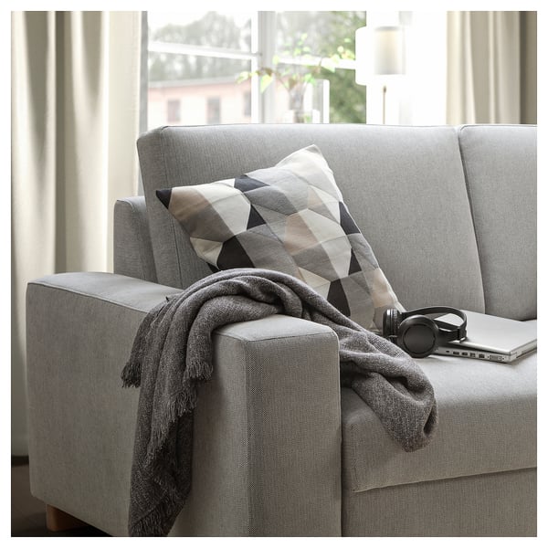 Modern grey sofa with geometric throw pillow and blanket, beside a window with a laptop and headphones.
