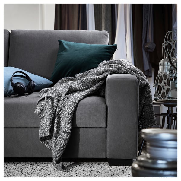 A modern grey SÖRVALLEN sofa with a green pillow and a cosy grey blanket. Headphones rest on it with a film reel nearby.