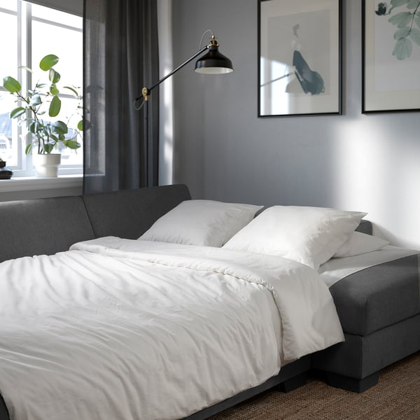 Modern sofa bed with white linens, suspended lamp, grey walls, and plant by window.