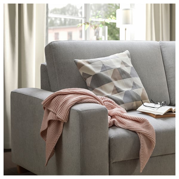 A light grey modern sofa with a cosy pink blanket and a patterned pillow sits near a window with beige curtains.