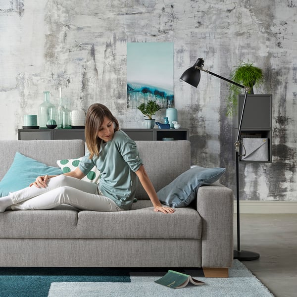 Person relaxes on grey SÖRVALLEN sofa, minimalist design, textured fabric, decorative throw pillow, modern living room with abstract art.