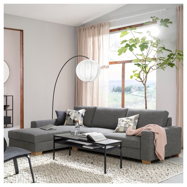 Modern grey l-shaped sofa with pillows, black coffee table, large plant, and hanging white light in bright living room.