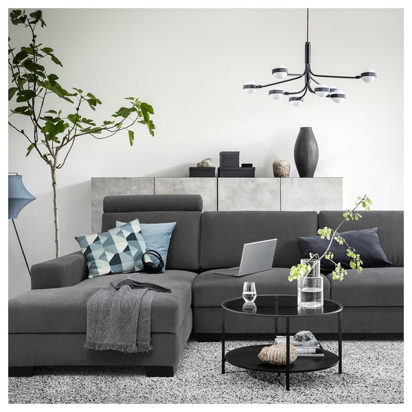Modern grey sectional sofa with patterned pillows, sleek coffee table, hanging lights, and a large plant in a minimalist living space.