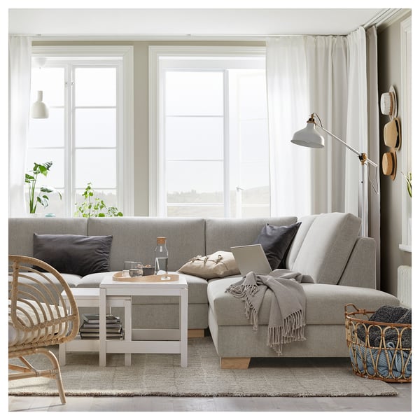 Cosy light grey sofa with multiple cushions and white coffee table, in a bright room with large windows and a floor basket with blankets.