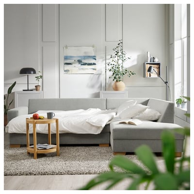 Modern living room with SÖRVALLEN light grey sofa-bed, white linens, wooden side table, large plants, and abstract wall art.