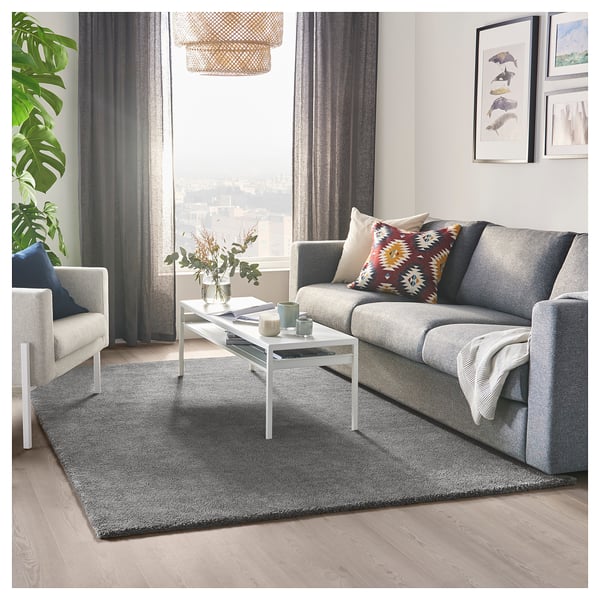 Modern living room with grey STOENSE rug, grey sofa, white coffee table, colourful throw pillows, and large windows with city view.