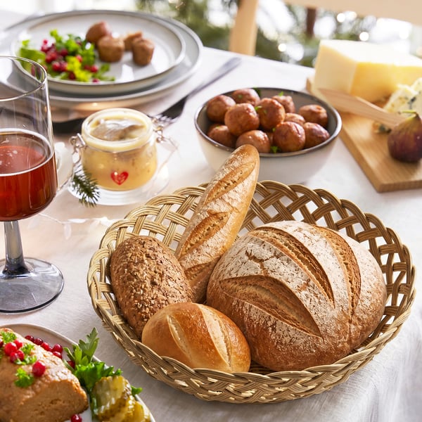 The STYLTSNÄPPA, a golden-rattan woven basket, sits on a table with bread. Large, holds fruit or bread, fits well with other dining items.