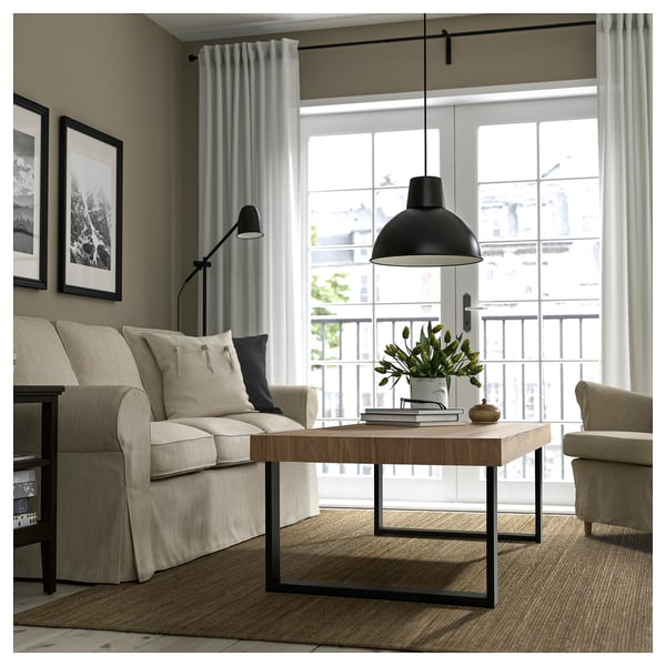 Modern living room with beige sofa, coffee table, floor lamp, and balcony access. Large window, curtains, and plant decor.