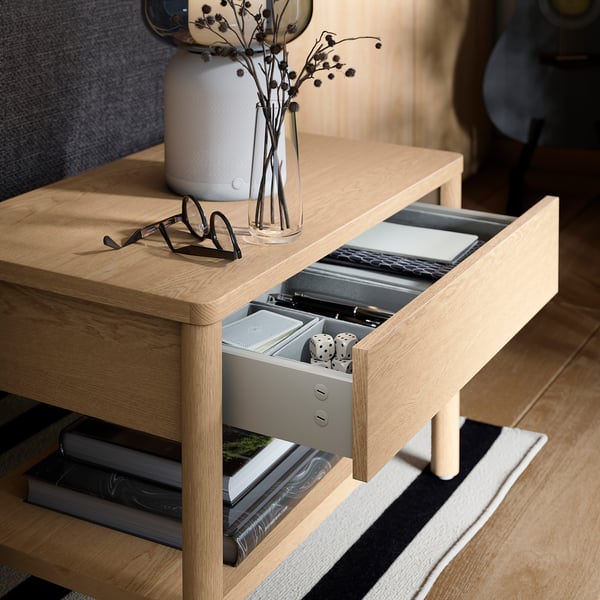 Wooden side table with open shelf and soft-close drawer. The drawer contains dice and a notebook. A vase with dried flowers and glasses are on top.
