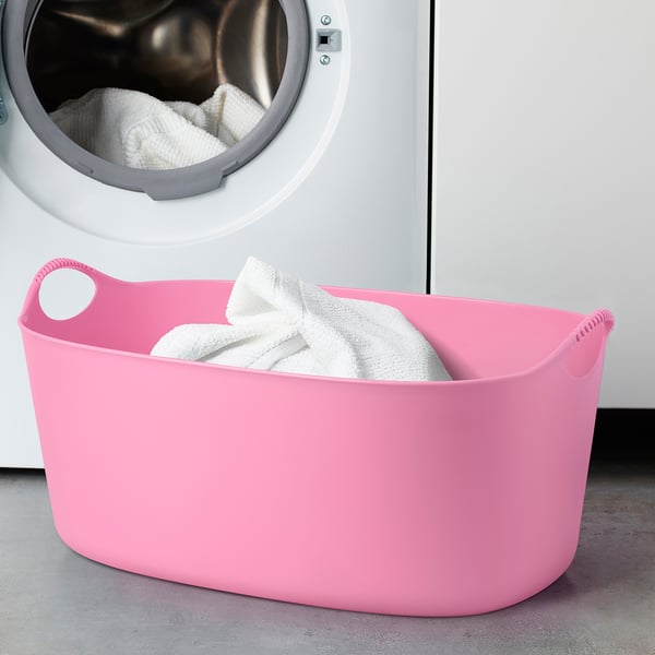 Bright pink TORKIS laundry basket with two handles, near white washing machine filled with white towels.