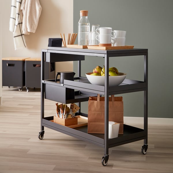 Modern metal three-shelf trolley with wheels, displaying mugs, apples, and bags.