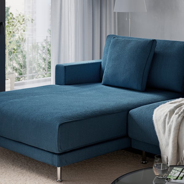 A blue l-shaped sofa with a pillow against the wall, near a lamp, window, and a table with a blanket and glass.