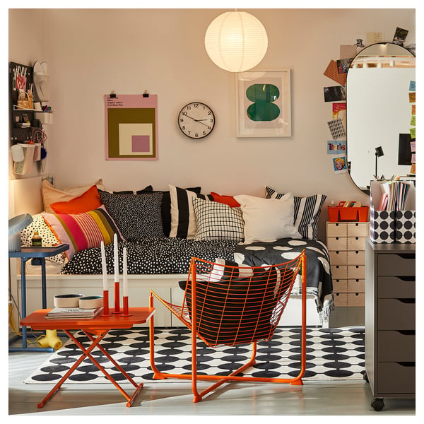 Bright bedroom with black dotted rug, colourful cushions, wall clock, hanging light, wall art, and orange furniture.