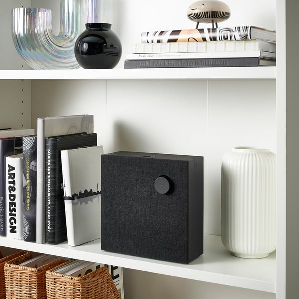 White bookshelf with a black speaker in the centre. Flanked by books, including jane packer living with flowers and a white vase.