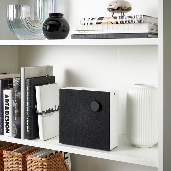 White bookshelf with books, a black vase, a unique speaker with a fabric cover, and a white textured vase.