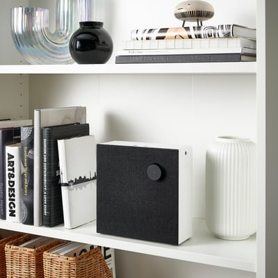 White bookshelf with books, a black vase, a unique speaker with a fabric cover, and a white textured vase.