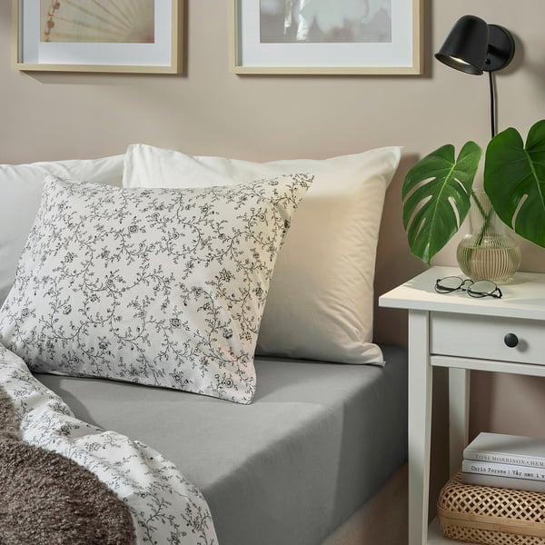 Cozy bed with VÅRVIAL sheets, patterned pillows. White side table with lamp, glasses, plant. Neatly made, minimalist decor.