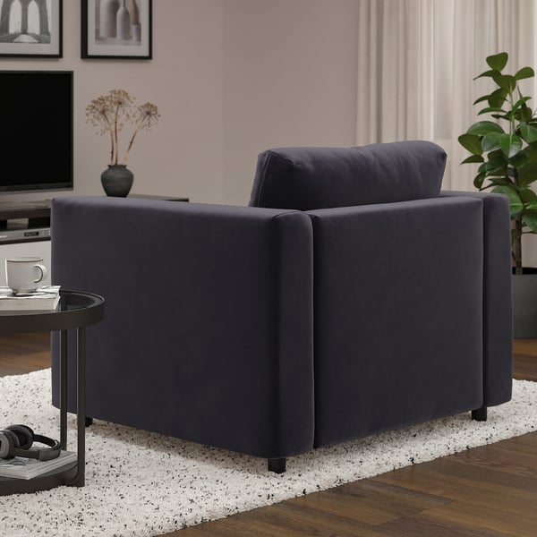 A cozy, dark grey armchair in a modern living space. Beside it, a small table holds a mug and headphones. Behind, a tv and a vase with dried flowers.