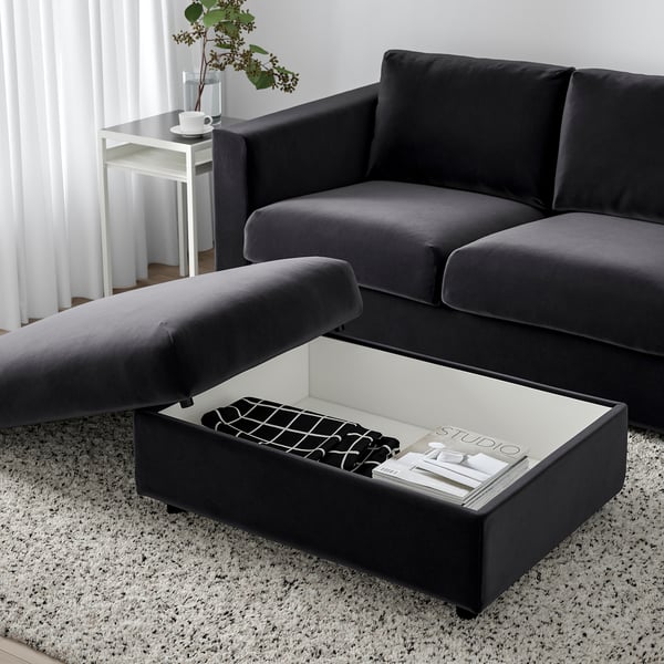 Dark grey VIMLE footstool with open storage, holding books and remote, beside matching sofa.