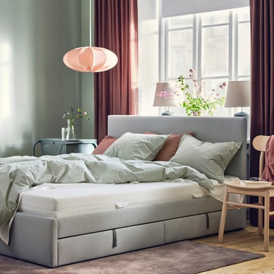Moderne soveværelse med lysegrå ÅBYGDA madras, hynder og hængelampe. Stort vindue med røde gardiner, natborde med lamper og planter.