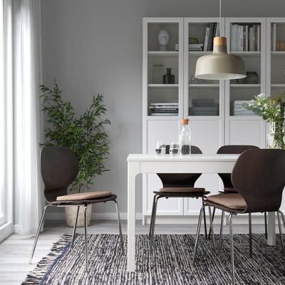 Moderne spisebordsopsætning med ÄLVGRÄSMAL stolepuder i beige, placeret på mørkebrune stole omkring et hvidt bord på et mønstret tæppe under en hængende lampe.