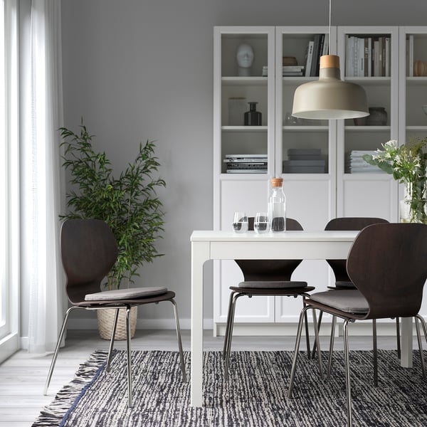 Moderne spisestue med grå puder, mørke stole, hvidt bord, bogreol, lys og plante.