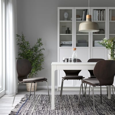 Moderne spisestue med grå puder, mørke stole, hvidt bord, bogreol, lys og plante.