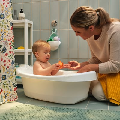 En kvinde bader en baby i et hvidt badekar ved hjælp af farverigt hvalformet legetøj.
