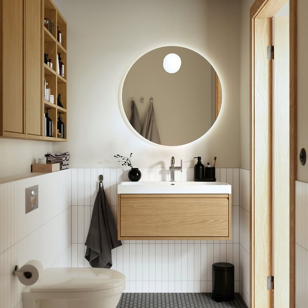 Moderne badeværelse: rundt spejl, egetræsvask, marmortop, sorte accessories, hvidt toilet, grå håndklæder.