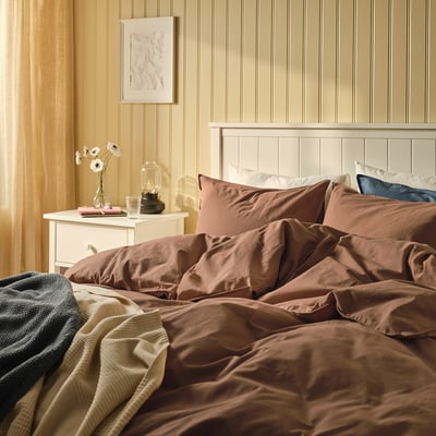 Hyggelig seng med brun ÄNGSLILJA-dyne , beige sengegavl, beige natbord med blomster.