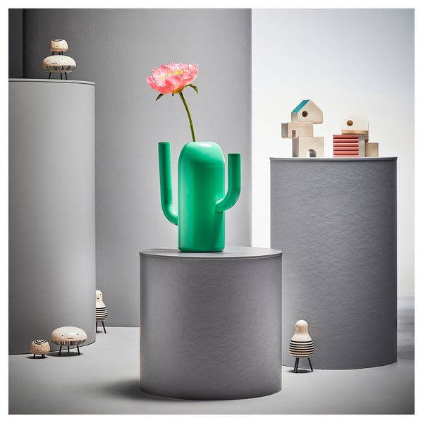 Lysegrøn kaktusformet vase på gråt stativ med blomster, omgivet af små dekorative figurer.