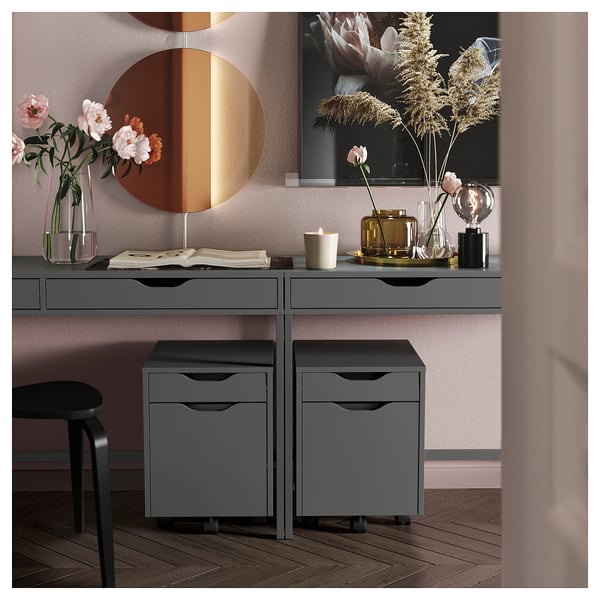 Moderne skrivebord med grå ALEX-skuffer, rundt spejl, blomster og dekorative genstande.
