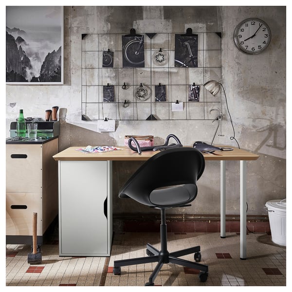 Skrivebordsopsætning i industriel stil med ANFALLARE bambusplade, sort stol, vægur og kunstudstilling.