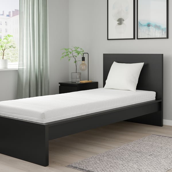 Minimalistisk soveværelse med ÅNNELAND sort enkeltseng, hvid pude, grønne gardiner, trægulv, sidebord med lampe og plante.
