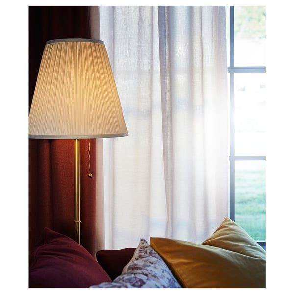 Hyggeligt opholdsrum med en rødbrun sofa, guldpuder og en høj lampe med en beige skærm og guldfod ved siden af hvide gardiner.