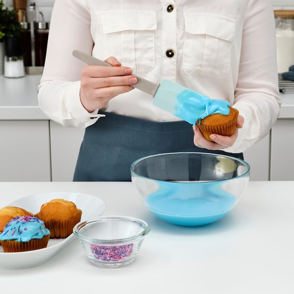 Person froster en muffin med en blå spatel ved hjælp af en klar skål til glasur.