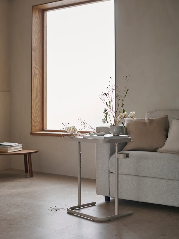 Moderne minimalistisk værelse med beige BJÖRKÅSEN laptopstativ ved stort vindue, grå sofa og træborde. Neutrale toner, rene linjer og naturligt lys.