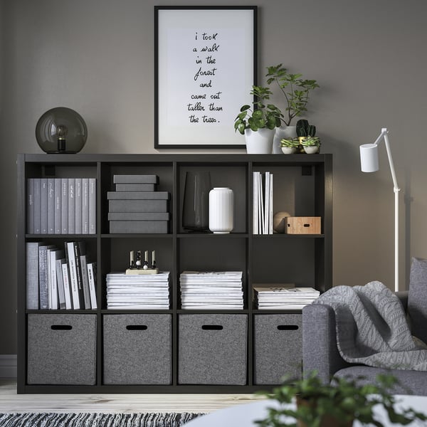 Sort kallax hylde med BLÄDDRA filt opbevaringsbokse, bøger, planter, indrammet kunst, lampe, grå sofa og gråt tæppe, organiseret og pænt.