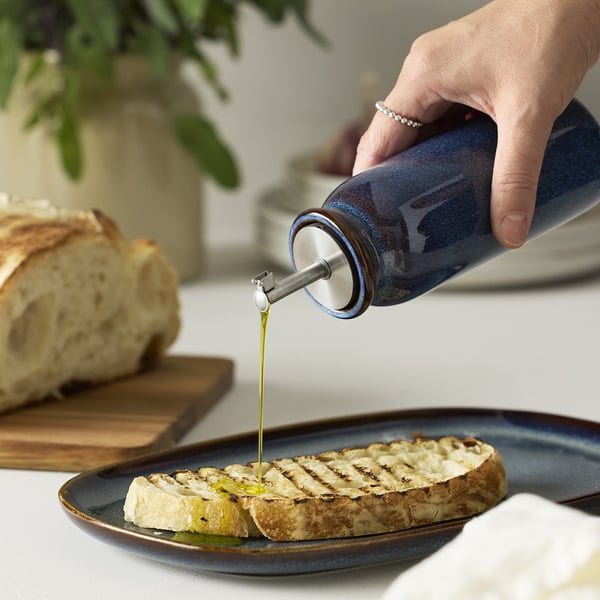 Håndhældning af olivenolie fra mørkeblå BRUGDHAJ-flaske på grillet brød på en matchende tallerken.
