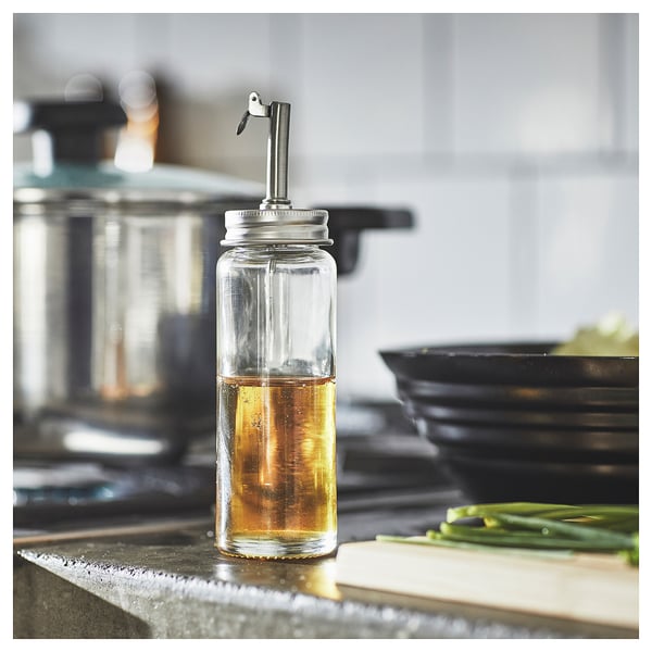 Oliedispenser i klart glas med metaldyse på køkkenbordet i nærheden af skærebræt og grønne løg. Enkelt, moderne design.