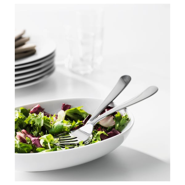 Hvid skål med grøn salat, to rustfri stålgafler hvilende på den, stak tallerkener og glas i baggrunden.