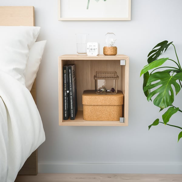 Væghængt EKET-terning i lyst træ, der rummer bøger, kasser, terrarium, ur og plante - minimal soveværelsesindretning.
