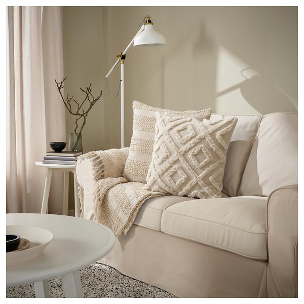 Beige sofa med hvide teksturerede puder, blød lampe, hvidt sidebord, bøger og dekorative grene.